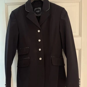 Svart ridkavaj med strass från Lime Cell - Snygg svart ridkavaj från Lime Cell med glittriga strassdetaljer på krage och fickor. Kavajen har silvriga knappar framtill och på ärmsluten, samt klassisk krage och ficklock. Perfekt för dig som vill sticka ut på tävling! Nypris 1500kr säljes för 650, endast använd två gånger! Så gott som ny🤩