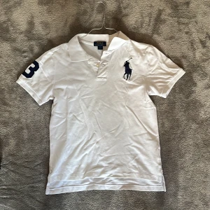 Vit piké från Polo Ralph Lauren - Vit pikétröja från Polo Ralph Lauren med klassisk krage och korta ärmar. Stor broderad logga på bröstet och siffran 3 på ena ärmen. Tröjan är i bomull och custom fit-passform. Storlek 14-16 år ungdom (passar på ungefär 160-170)