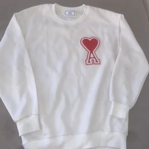 Vit sweatshirt från Ami med rött hjärta - !Läs hela beskrivningen!  Vit sweatshirt från Ami Alexandre Mattiussi med ett stort rött hjärta och bokstaven A broderat på bröstet. Tröjan har rund hals, ribbade muddar och är långärmad. Materialet är mjuk bomull och passformen är normal. Perfekt för dig som gillar stilrena detaljer med en twist. Tröjan har en liten gul fläck kolla bilderna
