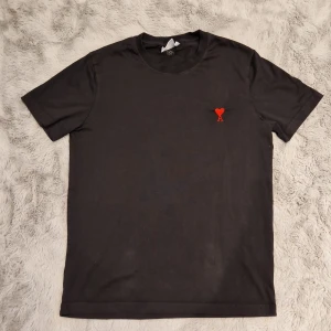 Svart t-shirt från Ami Alexandre Mattiussi - Svart t-shirt från Ami Alexandre Mattiussi i mjuk bomull. På bröstet finns ett broderat rött hjärta med bokstaven A, och baktill syns en diskret svart AMI-brodering. Klassisk passform och rund hals. Perfekt för dig som gillar stilrena detaljer. Priset kan diskuteras.