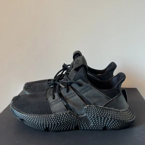 Svarta Adidas Prophere sneakers - Säljer ett par svarta Adidas Prophere sneakers med chunky sula och stickad ovandel. Skorna har snörning och detaljer i syntet och skinn. Riktigt coola och unika med sin robusta design och tydliga Adidas-logga på plösen.