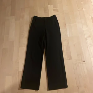 Svarta kostymbyxor från Vero Moda - Klassiska svarta kostymbyxor från Vero Moda med raka ben och midjehällor. Byxorna har dragkedja och knapp framtill.