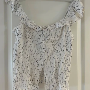 Vit blommig topp med volang H&M - Supersöt vit blus från H&M med svart blommigt mönster, volang runt axlarna och knappar framtill. Blusen har en lätt och luftig känsla, perfekt för varmare dagar. Den är ärmlös och har en liten slits framtill för extra detalj. pris kan diskuteras, hör av er vid frågor!💗