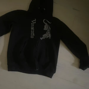 Svart Von Studio hoodie Large - Svart hoodie från Von Studio med dragkedja, huva och långa ärmar. Framsidan har ett glittrigt strassmönster med en tiger och Von Studio-loggan. Perfekt för dig som gillar streetwear med lite extra bling. Materialet känns som mjuk bomull och polyester. Den är i storlek Large