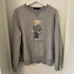 Grå sweatshirt Polo Bear Ralph Lauren - Grå sweatshirt från Polo Ralph Lauren med ikoniska Polo Bear-trycket framtill. Klassisk rund halsringning, ribbade muddar och avslappnad passform. Perfekt för dig som gillar stilrena och lekfulla detaljer. Materialet är mjuk bomullsmix.