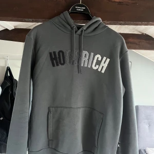 Grå HOODRICH hoodie med brodyr - Snygg grå hoodie från HOODRICH med broderad logga i svart, grått och vitt framtill. Klassisk känguruficka, huva med snörning och långa ärmar. Perfekt för dig som gillar streetwear och vill ha en stilren look.