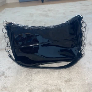 Svart axelväska med kedjedetaljer - Snygg svart axelväska i glansigt syntetmaterial med silvriga kedjor på sidorna och dragkedja upptill. Perfekt storlek för det viktigaste och ger en cool vibe till din outfit.