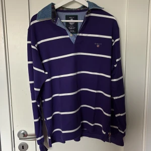 Randig lila pikétröja från Gant - Säljer en lila och vitrandig pikétröja från Gant med denimkrage och klassisk logga på bröstet. Tröjan har långa ärmar och relaxed fit, perfekt för en chill och stilren look. Materialet är mjuk bomull och kragen har snygga kontrastdetaljer.