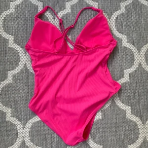 Ribbad rosa baddräkt med snörning - Snygg ribbad baddräkt i stark rosa färg med smala axelband och snörning fram. Modellen har en djup v-ringning och är tillverkad i mjuk och stretchig polyester som sitter skönt mot kroppen. Perfekt för strandhäng och poolparty i sommar!