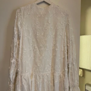 Vit blommig kortklänning med öppen rygg - Superfin vit kortklänning med blommigt broderat mönster och volangdetaljer. Klänningen har långärmade ärmar och knyt i nacken. Perfekt för sommarens alla tillfällen, med romantisk och luftig vibe. Materialet känns som bomull och ger en skön känsla mot huden.