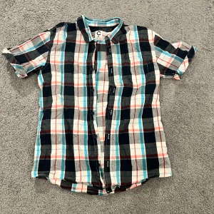 Rutig kortärmad skjorta från Cubus - Kortärmad skjorta i bomull med rutigt mönster i svart, grått, vitt, rött och blått. Klassisk krage, knäppning framtill och bröstficka. Perfekt för en avslappnad och cool stil.