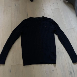 Mörkblå stickad tröja från GANT - Säljer en mörkblå stickad tröja från GANT i storlek L. Tröjan har ett klassiskt kabelstickat mönster och rund hals. Perfekt för dig som gillar stilrena och tidlösa plagg. GANT-logga broderad på bröstet. Långärmad och mjuk kvalitet.