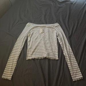 Randig offshoulder topp Kappahl - Superfin vit offshoulder topp från Kappahl med svarta tunna ränder. Långärmad modell med croppad passform och volangkant nertill och vid ärmslut. Perfekt för dig som gillar en enkel men ändå söt stil.