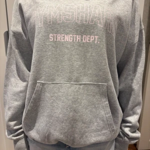 Gymshark hoodie - Säljer en grå hoodie från Gymshark med ljusrosa text 'Strength Dept.' framtill. Tröjan har huva, magficka och är långärmad. Oversized passform och mjukt material, perfekt för chill eller träning. Snygg och enkel att matcha med jeans eller tights.