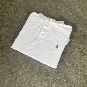 Vit pikétröja från Ralph Lauren - Vit klassisk pikétröja från Polo Ralph Lauren i bomull. Tröjan har korta ärmar, krage och knäppning framtill samt den ikoniska broderade loggan på bröstet. Perfekt för en clean och stilren look.