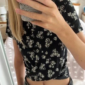 Svart topp från Gina Tricot - Supersöt svart kort topp från Gina Tricot med vita blommor över hela. Modellen är croppad med en twistad detalj framtill och har korta ärmar. Knappt använd, så inga defekter. Den är i storlek XS, hör av er om frågor!❤️