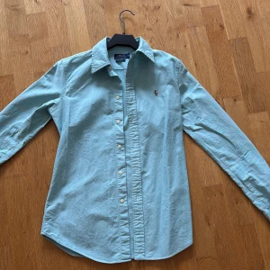 Ljusblå skjorta från Polo Ralph Lauren - Klassisk ljusblå skjorta från Polo Ralph Lauren med broderad logga på bröstet. Skjortan har lång ärm, knappar framtill och en snygg krage. Perfekt för en stilren och avslappnad look.