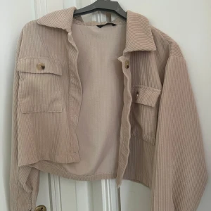 Beige manchester overshirt - Snygg beige overshirt i manchester med krage och två stora bröstfickor med knappar. Jackan har en avslappnad passform och är perfekt att slänga över en t-shirt. Ribbade detaljer och croppad längd ger en trendig vibe. Använt några gånger.