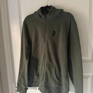 Olivgrön hoodie Peak Performance XL - Olivgrön hoodie från Peak Performance i storlek XL. Tröjan har dragkedja framtill, två fickor och en skön huva. Tillverkad i mjuk bomullsblandning, perfekt för chill dagar. Diskret logga på bröstet och klassisk passform.