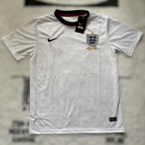 England fotbollströja Nike 150 år - England fotbollströja från Nike, officiell jubileumsmodell för 150 år. Vit med mörkblå krage, broderat landslagsmärke och guldiga detaljer. Tillverkad i lätt och ventilerande Dri-FIT-material, perfekt för match eller träning.