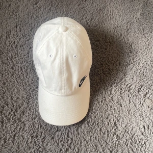Vit keps från Nike med broderad logo - Snygg vit keps från Nike med klassisk böjd skärm och svart broderad Nike-logo framtill. Justerbar rem baktill för perfekt passform. Tillverkad i bomull, perfekt för en clean och sportig look.