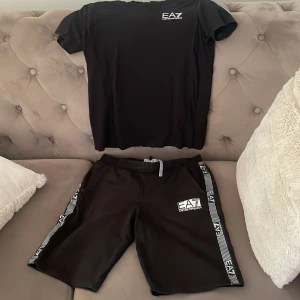 Svarta EA7 shorts med logga - Snygga svarta shorts från EA7 Emporio Armani med vit logga framtill och coola ränder med EA7-tryck längs sidorna. De har midwaist passform, snörning i midjan och är gjorda i mjukt syntetmaterial. Perfekta för chill eller sportiga dagar.
