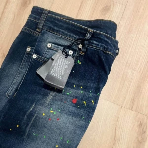 Dsquared2 jeans med färgstänk - Mörkblå jeans från Dsquared2 med coola färgstänk i gult, grönt och rött. Klassisk femficksmodell med slitningar och en grön Dsquared2-patch bak. Jeansen har en rak passform och är tillverkade i bomullsdenim.