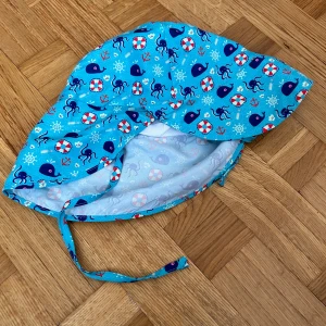Blå solhatt med havsmotiv och knytband - Söt blå solhatt med knytband under hakan och marint mönster med valar, bläckfiskar, livbojar och ankare. Hatten har bred skärm för extra solskydd och är tillverkad i mjuk bomull, perfekt för soliga dagar.