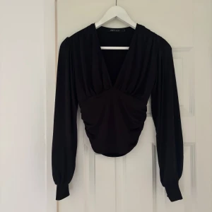 Svart v-ringad topp från NLY One - Svart långärmad topp från NLY One med snygg v-ringning och rynkade detaljer framtill. Modellen har puffiga ärmar med smala muddar. Perfekt för dig som gillar stilrena och trendiga plagg.