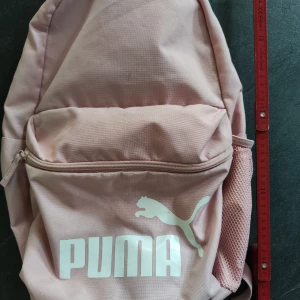 Ljusrosa ryggsäck från Puma - Säljer en ljusrosa ryggsäck från Puma med stort vitt logotryck framtill. Väskan har ett stort huvudfack, en framficka med dragkedja och meshficka på sidan. Justerbara, vadderade axelremmar för extra komfort. Perfekt för skola eller cityhäng.