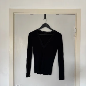 Svart ribbad topp med spets från BikBok - Svart långärmad topp från BikBok med ribbad struktur och djup v-ringning dekorerad med spetsdetaljer. Toppens kanter har en lätt vågig finish och den är figurnära i modellen. Perfekt för dig som gillar en stilren men ändå söt look.