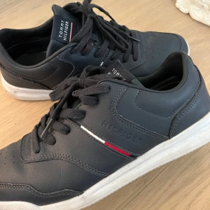 Mörkblå sneakers från Tommy Hilfiger - Snygga mörkblå sneakers från Tommy Hilfiger med klassisk logga på sidan och bak. Skorna har vita och röda detaljer, svart snörning och vit sula. Skornas original pris är 2100kr!! Tillverkade i skinn och syntet, perfekt för en stilren och sportig look.