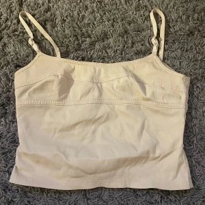 Beige linnetopp med smala axelband - Snygg beige linnetopp med justerbara smala axelband och sömdetaljer över bysten. Toppen är croppad och har en enkel, stilren design som passar perfekt till jeans eller kjol. Gjord i mjukt bomullstyg för en skön känsla. Är i storlek 38 och kommer ifrån H&M.