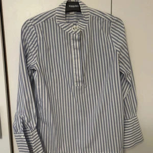 Randig blåvit henley från KappAhl - Snygg henley-skjorta från KappAhl i blåvitt randigt mönster. Skjortan har lång ärm, ståkrage med dekorativ knapp och slits vid ärmslut. Tillverkad i mjuk bomull som känns skön mot huden. Perfekt för dig som gillar en clean och stilren look.