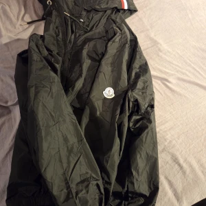 Svart vindjacka från Moncler - Snygg svart vindjacka från Moncler med klassisk logga på armen. Jackan har huva  och dragkedja framtill. Lätt och smidig modell som passar perfekt när det blåser. Detalj med röd och vit rand på huvan. Är runt 198cm och den va lite liten. NFC scan funkar