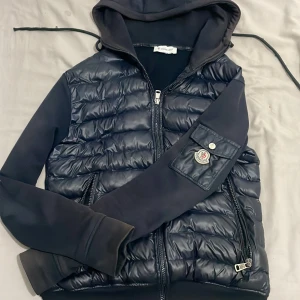 Svart pufferjacka med huva från Moncler - Snygg svart pufferjacka från Moncler med huva och dragkedja. Jackan har quiltade partier framtill och på ärmen finns Moncler-logga och ficka med knapp. Materialmix med glansig polyester och mjuka partier. Perfekt för dig som vill ha en stilren och sportig look. Jackan har nfc.  Priset kan diskuteras vid snabbäffär