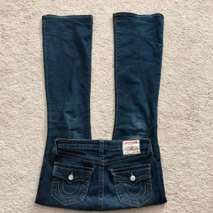 True Religion Becca Bootcut Jeans - Mörkblå jeans från True Religion, modell Becca Mid Rise Bootcut. Klassisk femficksdesign med snygga kontrastsömmar och ikoniska bakfickor med lock och knapp. Jeansen har medelhög midja och bootcut-ben, tillverkade i slitstarkt denim.