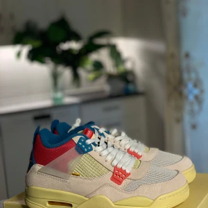 Nike Air Jordan 4 Union Guava Ice - Nike Air Jordan 4 Union Guava Ice med en mix av beige mocka, mesh och färgglada detaljer i rött, blått och gult. Snygg retro-vibe med vit snörning, synliga plastdetaljer och ikonisk Nike Air-logga på hälen. Sulan är gul med röda och blå accenter undertill.