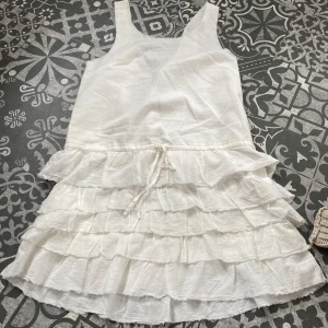 Vit volangklänning med knyt - Supersöt vit kortklänning med flera lager volanger och knyt i midjan. Ärmlös modell i lätt och luftig bomull, perfekt för varma dagar. Aldrig använd! ☀️