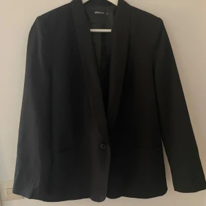 Svart kavaj från Gina Tricot, stl 38 - Stilren svart kavaj från Gina Tricot i storlek 38. Klassisk modell med en knapp framtill, två knappar vid ärmslut och slät krage. Perfekt för en clean och snygg look. Tunt fodrad och tillverkad i ett mjukt material.