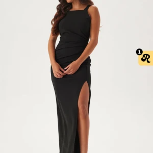 Svart långklänning med slits - Supersnygg svart långklänning med breda axelband, figurnära passform och hög slits på ena sidan. Klänningen har draperade detaljer i midjan och en öppen rygg med dragkedja. Perfekt för dig som vill sticka ut med en elegant och stilren look. Endast använd 2 gånger