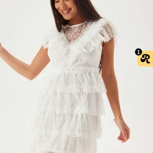 Vit spetsklänning med volanger bubbelroom - Superfin vit kort spetsklänning från bubbleroom med flera lager volanger, v-ringning och transparent spets upptill. Klänningen har korta ärmar och en romantisk känsla med snygga detaljer. Perfekt för dig som vill sticka ut med en söt och trendig look.