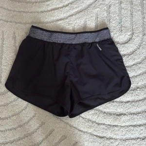 Svarta träningsshorts Domyos XS - Svarta träningsshorts från Domyos med elastisk, mönstrad midja i grått. Lätta och luftiga i 100% polyester, perfekta för gym eller löpning. Snygg sportig look och bekväm passform. Märke och storlek tydligt markerade.