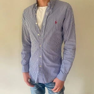 Randig skjorta från Ralph Lauren - Snygg blå- och vitrandig skjorta från Ralph Lauren Skjortan har lång ärm och den ikoniska röda loggan broderad på bröstet. Perfekt för en stilren och avslappnad look. Skriv vid, minsta fråga!
