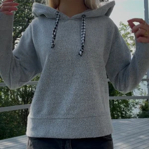 Grå hoodie med rutiga snören - Mysig grå hoodie med fluffig känsla och svartvitt rutiga snören i luvan. Tröjan har lång ärm, relaxed fit och stor huva. 