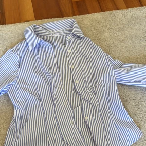Blåvit randig skjorta med knappar - Snygg skjorta med blå och vita ränder och klassisk krage. Skjortan har långa ärmar och stängs med vita knappar framtill. Perfekt för en clean och fräsch look. Tillverkad i mjuk bomull som känns skön mot huden.