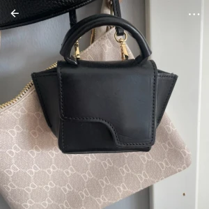 mini APT väska  - Säljer en svart minihandväska i skinn med stilren design och guldfärgade detaljer. Väskan har ett litet handtag och en unik form som sticker ut. Perfekt för dig som gillar trendiga accessoarer och vill ha något som syns. Säljer min mini atp väska som är söt o liten!