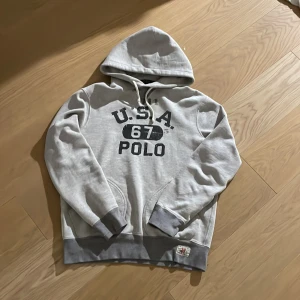 Grå hoodie från Polo Ralph Lauren - Snygg grå hoodie från Polo Ralph Lauren med tryck 'U.S.A. 67 POLO' på bröstet. Storlek M men passar även S. Tröjan har huva med snörning, känguruficka och ribbade muddar. Perfekt för en avslappnad och sportig stil. Inga defekter. Hör av vid minsta fundering. 💯💯