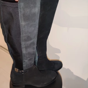 Svarta mockastövlar Tommy Hilfiger - Snygga svarta stövlar från Tommy Hilfiger i mocka med stretchig textil på skaftet. Dekorativt metallspänne vid hälen och dragkedja på insidan. Fodret har coolt mönster med färgglada bokstäver. Perfekta för höst och vinter!