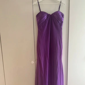 Lila långklänning - Säljer en lila långklänning med tunna axelband och vackert draperad överdel. Klänningen har en elegant v-ringning och är tillverkad i ett glansigt material som ger en lyxig känsla. Perfekt för bal eller festliga tillfällen. Funkar att använda utan axelbandet. Tyvärr har klänningen små defekter fram till